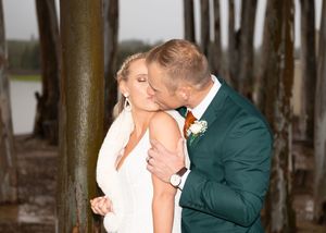 A romantic wedding couple shares an intimate kiss between wooden pier posts at sunset.