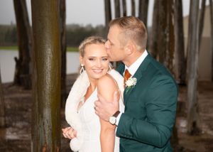 A couple in wedding attire share a romantic moment among trees, with the bride in white and groom in emerald green.
