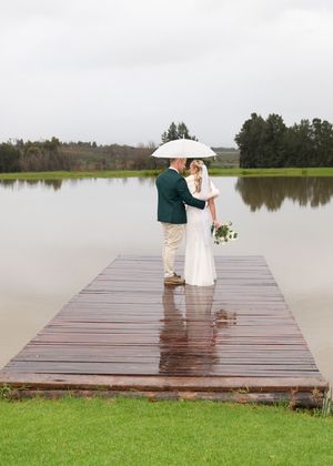 A couple shares an intimate moment under a white umbrella on a wet wooden dock overlooking a serene lake.