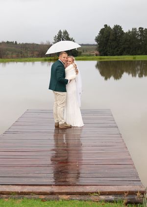 A couple stands close together on a wet wooden dock over a lake while sharing a white umbrella on an overcast day.