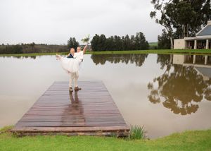 A figure in a flowing white dress stands on a wooden dock overlooking a serene lake with trees and a building in the background.