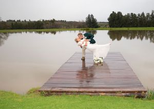 A couple embraces on a wooden dock overlooking a serene lake on an overcast day with green grass in the foreground.