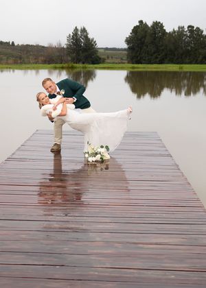 A romantic wedding photo captured on a wet wooden dock over a serene lake with trees in the background and floral bouquet.
