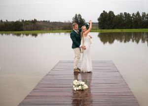 A couple celebrates joyfully on a wooden dock overlooking a serene lake, with a bouquet of flowers at their feet.