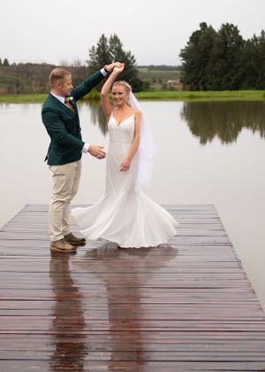 A couple dances joyfully on a wet wooden dock overlooking a serene lake on their wedding day.