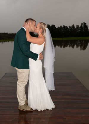 A couple shares their first kiss as newlyweds on a wooden dock by a tranquil lake at dusk.