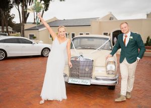 A vintage silver car parked on a brick driveway with two people celebrating in formal attire.