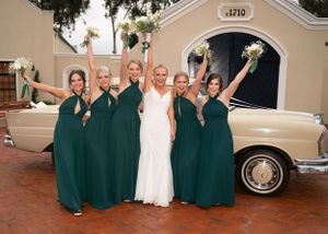 Wedding party in emerald green bridesmaid dresses pose with bride in white gown next to vintage cream-colored car.