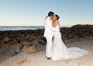 A romantic wedding couple embraces on a rocky beach at sunset with the ocean and coastline in the background.