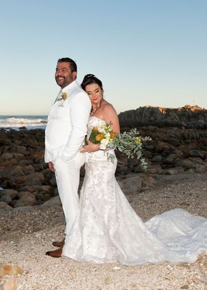 A couple in white wedding attire poses on a rocky beach at sunset with mountains and ocean in the background.