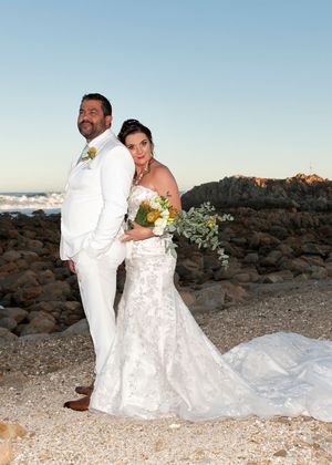 A newlywed couple poses on a rocky beach at sunset, with the bride in white lace and groom in a white suit.