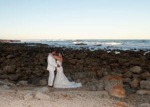A couple in wedding attire embrace on a rocky beach at sunset with waves crashing in the background.