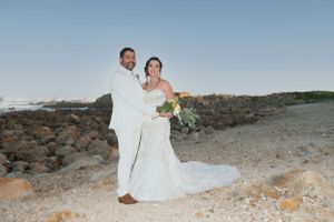 A couple in wedding attire stands together against a rocky coastal backdrop at sunset.