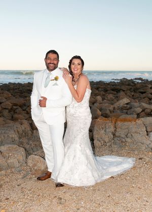 A couple wearing white attire poses for their wedding photo on a rocky beach at sunset.