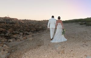 A couple in wedding attire walks along a rocky coastal path during sunset.