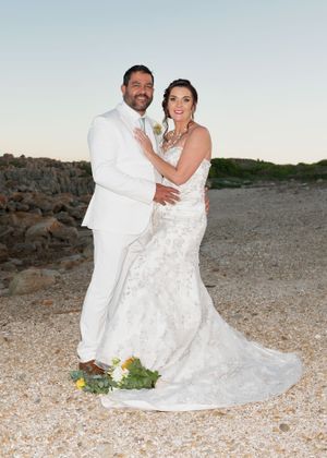 A couple in matching white attire poses for a wedding photo on a beach at sunset with a rocky coastline in the background.