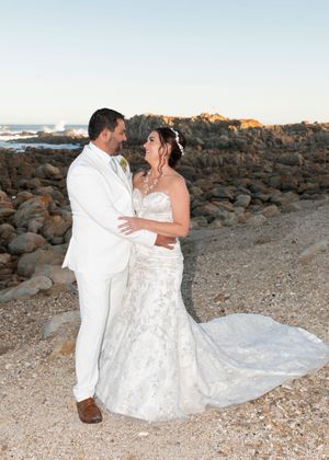A couple embraces on a rocky beach with ocean waves and golden sunset light in the background.