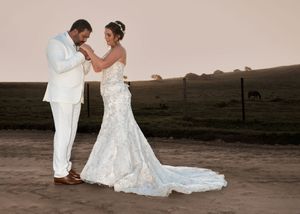 A couple in elegant white wedding attire standing together at sunset with rolling hills in the background.
