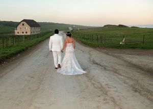 A couple in wedding attire walk down a country road at sunset with a barn and open fields in the distance.