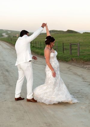 A couple in white wedding attire dance together on a dirt road surrounded by rolling green hills and fencing.