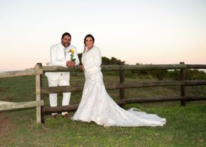 Newlyweds pose together by a rustic wooden fence on their wedding day with scenic views in the background.