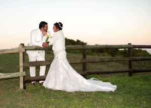 A couple in wedding attire share a romantic moment leaning against a wooden fence at sunset by the water.