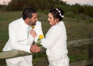 A romantic wedding couple in matching white outfits share a drink together while leaning on a wooden fence at sunset.