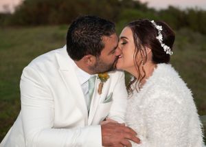 A couple in white wedding attire share a romantic kiss outdoors during their wedding ceremony.