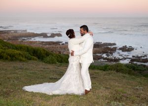 A couple in wedding attire embraces on a scenic coastal cliff at sunset overlooking rocky shores and ocean waves.