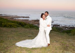 Couple in white wedding attire embrace on coastal cliff during sunset with ocean waves crashing on rocky shore below.