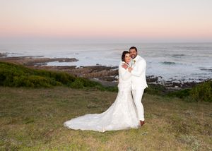 A couple in wedding attire embraces on a cliff overlooking a scenic ocean coastline at sunset.