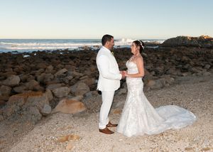 A couple in elegant white wedding attire stands together on a rocky beach shoreline at sunset.