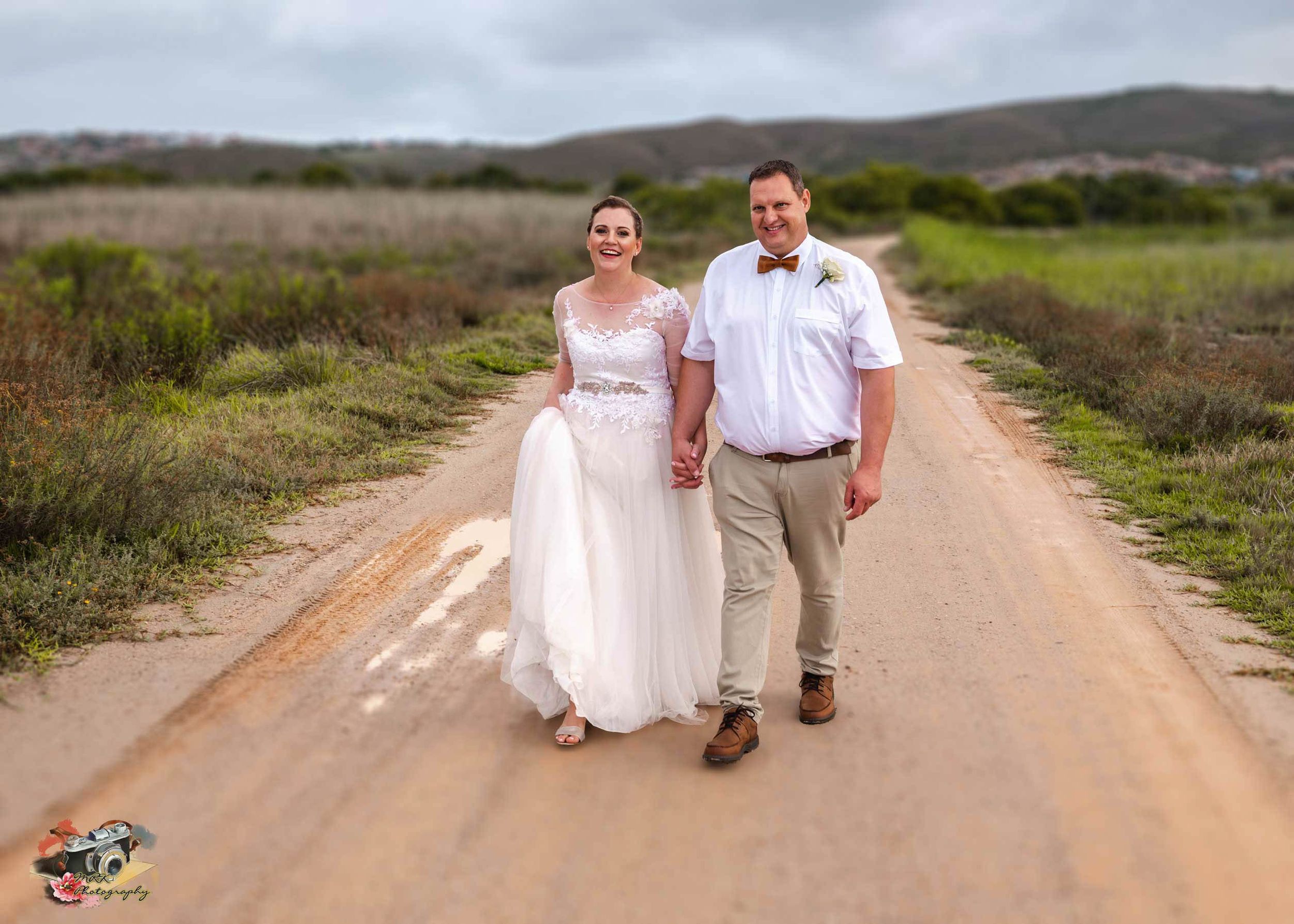 A couple walks hand in hand on a dirt road through countryside scenery wearing wedding attire on a sunny day.