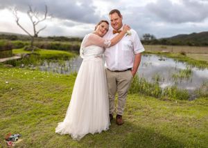 A rustic outdoor wedding photo with a couple embracing on grassy hills overlooking a scenic pond under cloudy skies.