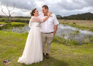 A happy couple celebrates their wedding day outdoors near a pond with rolling hills and cloudy skies in the background.