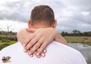 Hand with engagement ring rests on person's shoulder with a pond and cloudy sky in the background.
