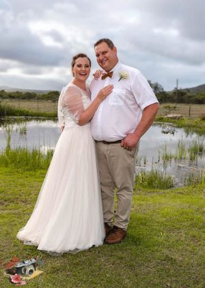 Newlywed couple poses together outdoors by a pond on their wedding day under cloudy skies.