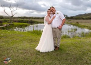 A romantic wedding couple embraces near a tranquil pond on a cloudy day with green grass and wetlands in the background.