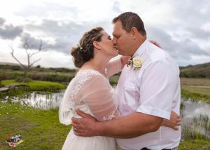 A romantic couple shares a tender kiss outdoors by a pond with cloudy skies and rolling hills in the background.