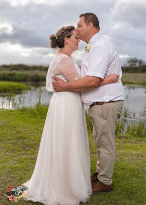A couple in wedding attire share a romantic moment beside a marshy pond under cloudy skies.