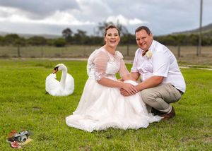 A couple in wedding attire sits in a grassy field while a white swan approaches in the background.