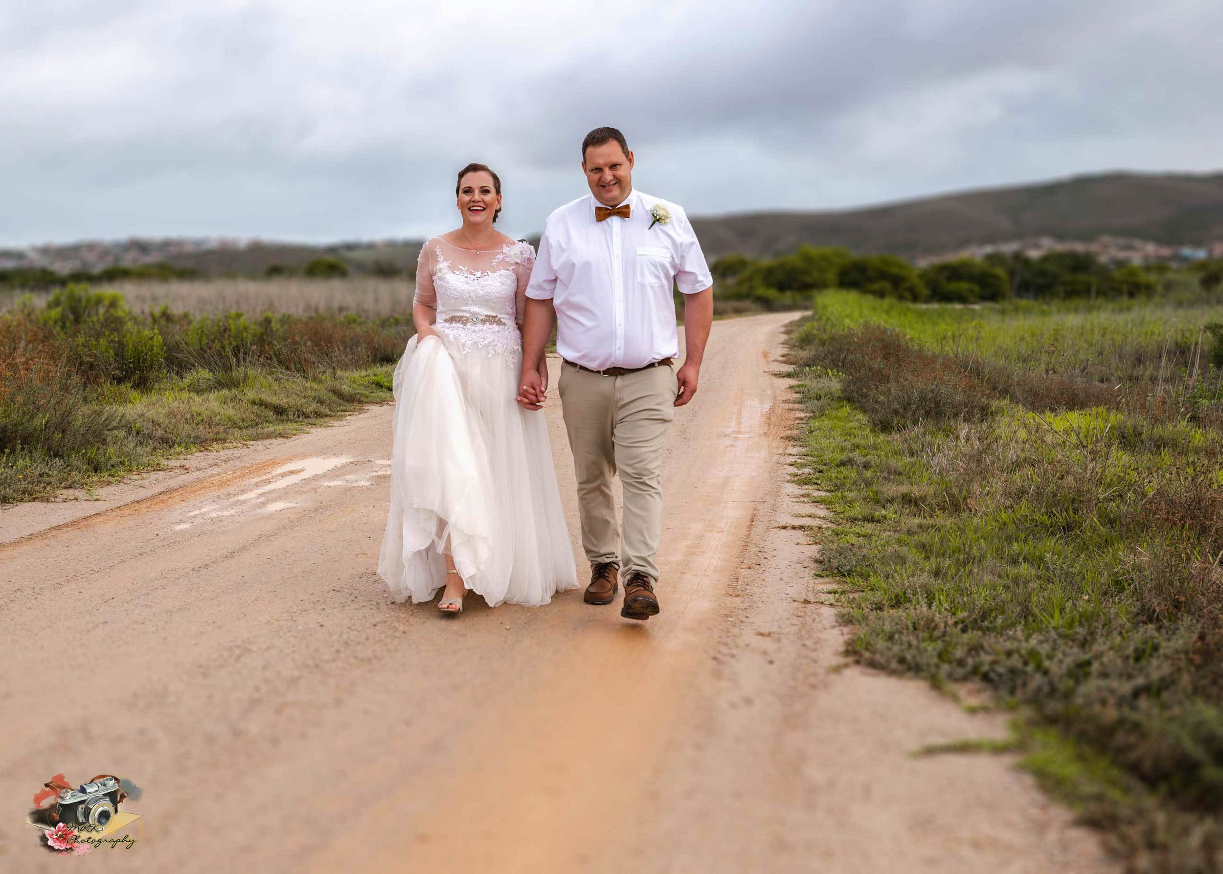 Newly married couple walking together down a dirt road in rustic countryside setting with hills in background.