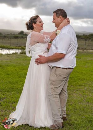 A wedding couple embraces and laughs together outdoors on a green field under dramatic cloudy skies.