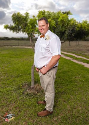 Someone in a white shirt and khaki pants poses outdoors with a bowtie and boutonniere on a grassy field.