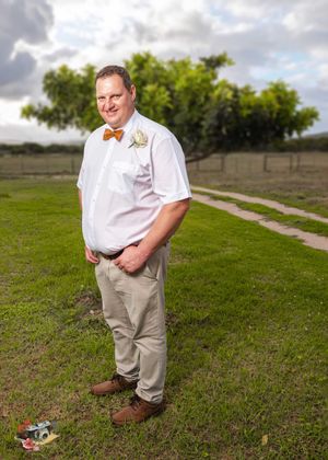 A person wearing casual wedding attire stands on grass with trees in the background under cloudy skies.