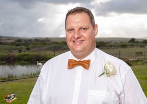 Wedding photo of person in white dress shirt and brown bow tie against scenic landscape with water and cloudy sky backdrop.