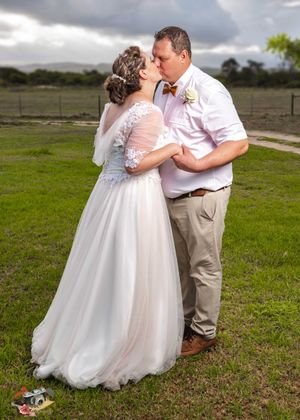 A romantic wedding couple share a tender kiss outdoors on a grassy field with cloudy skies in the background.
