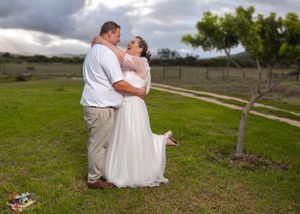 A romantic wedding couple shares a joyful embrace on green countryside with cloudy skies in the background.