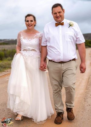 A newlywed couple poses on a dirt road in casual wedding attire with khaki pants and a bow tie against a cloudy sky.