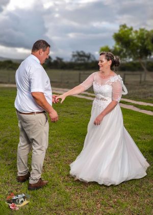 A couple dancing together outdoors on grass with cloudy skies, the bride wearing a flowing white lace wedding gown.
